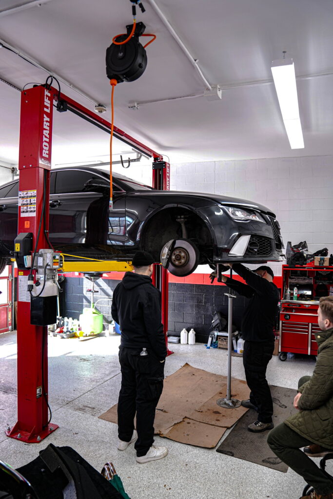 Two mechanics from W12 Garage inspecting an Audi sedan's struts and suspension.